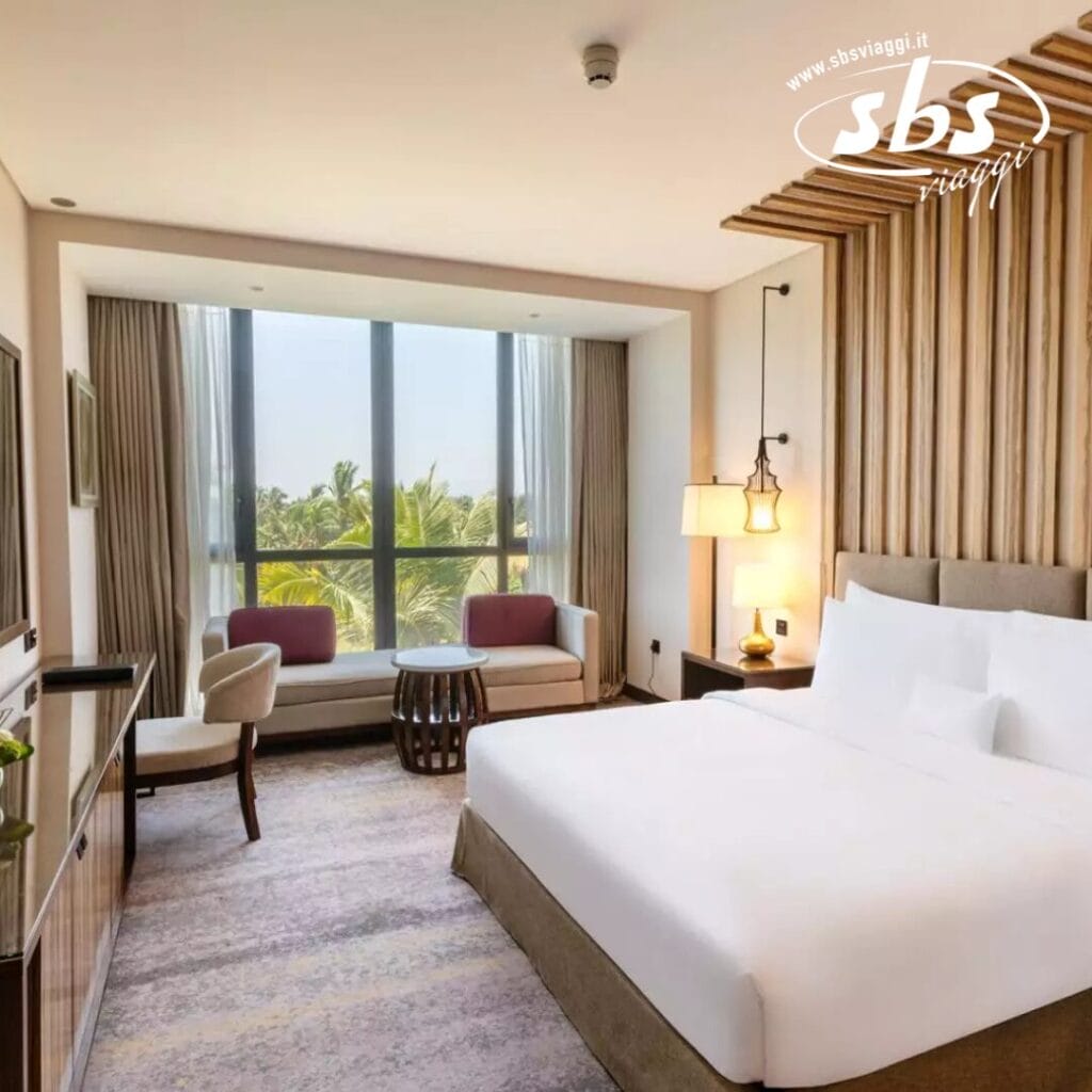 L'interno della camera d'albergo è caratterizzato da un letto grande e da un'accogliente zona salotto vicino alla finestra con tende. Una TV è elegantemente appoggiata sul suo supporto, circondata da un arredamento moderno. Il logo 