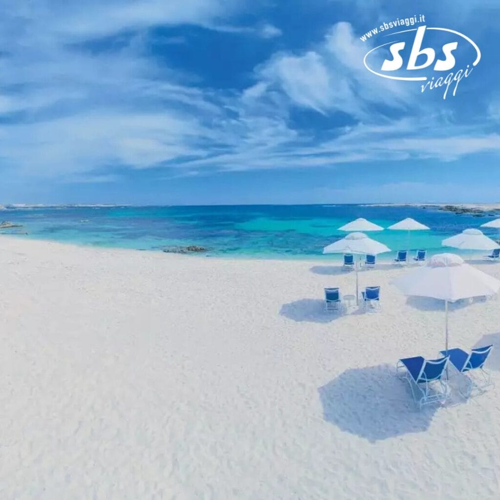 Una spiaggia incontaminata con sabbia bianca e acqua azzurra presenta diverse sedie a sdraio blu sotto ombrelloni bianchi. Il logo "sbs viaggi" è ben visibile, aggiungendo un tocco elegante a questa fuga idilliaca.