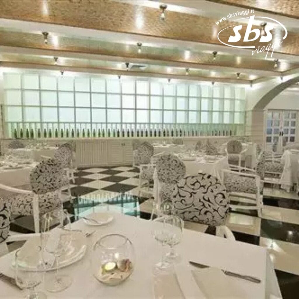 L'elegante interno del ristorante è caratterizzato da sedie a fantasia e tovaglie bianche su un pavimento a scacchiera. I tavoli sono apparecchiati con bicchieri e piatti, mentre un espositore di bottiglie di vino decora la parete di fondo, aggiungendo un tocco di bozza automatica all'ambiente.