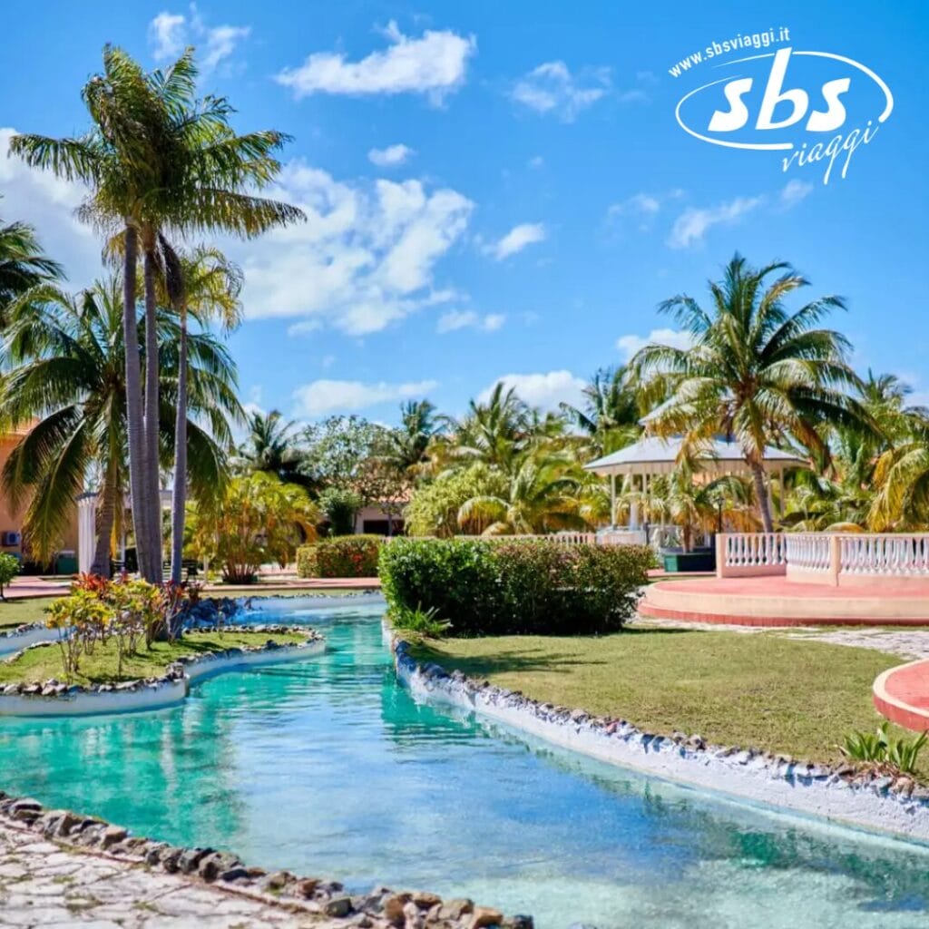 Scena di un resort tropicale con una piscina tortuosa, palme e un gazebo sotto un cielo azzurro terso. Il logo