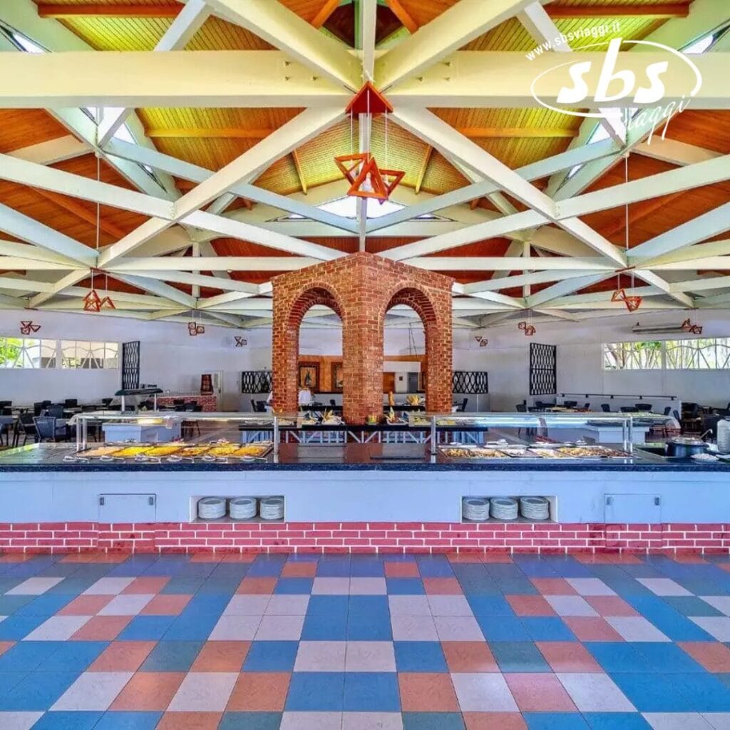 Una spaziosa sala da pranzo nel resort Veraclub accoglie gli ospiti con un grande tavolo da buffet al centro. Il soffitto è caratterizzato da una struttura in legno con travi a vista, mentre il pavimento è splendidamente piastrellato con motivi blu e rosa, che ricordano il fascino vibrante di Varadero.