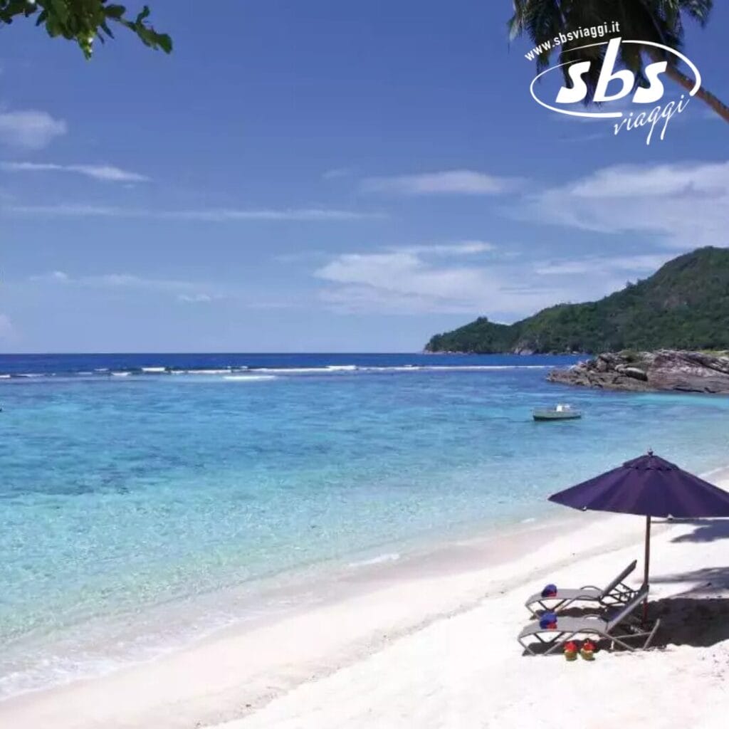 Si apre una scena di spiaggia serena con un mare calmo e cieli limpidi. Una sedia a sdraio vuota sotto un ombrellone blu invita al relax sulla sabbia, evocando una tranquillità da spa. In lontananza, le colline incorniciano lo sfondo, ricordando i tranquilli dintorni dell'Hilton Allamanda Resort.