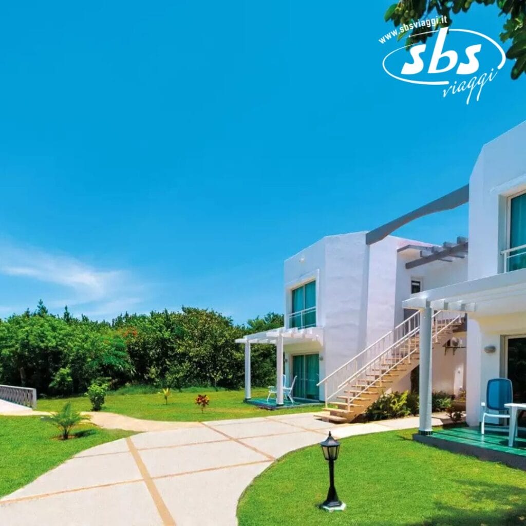 Un edificio bianco a due piani con balconi si erge in mezzo a un prato verde lussureggiante e alberi sotto un cielo azzurro e terso. Un sentiero conduce all'ingresso, dove il logo