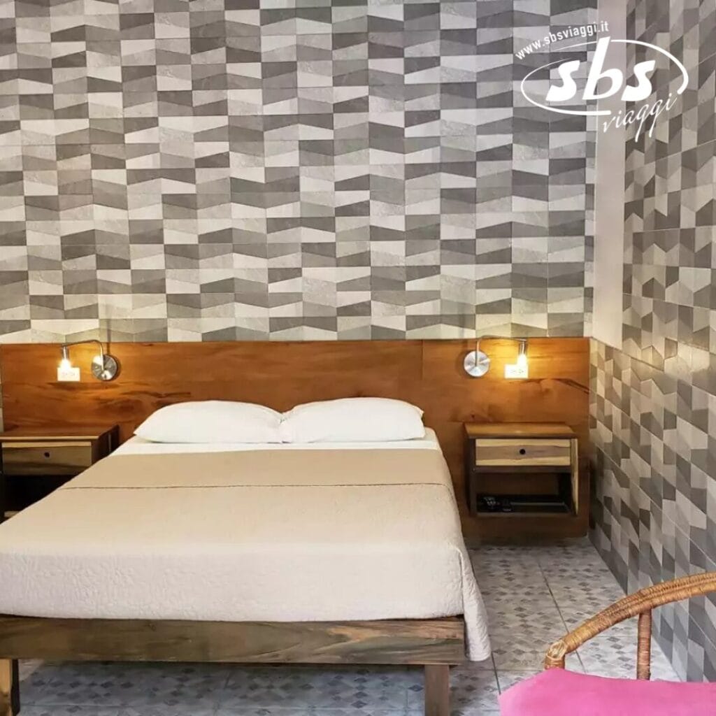 Una camera da letto in stile Sunrise Club presenta un letto matrimoniale contro una parete con motivi geometrici, con luci a parete e comodini su entrambi i lati. Una sedia rosa è sistemata con grazia nell'angolo, aggiungendo un tocco di colore a questo rifugio sereno.