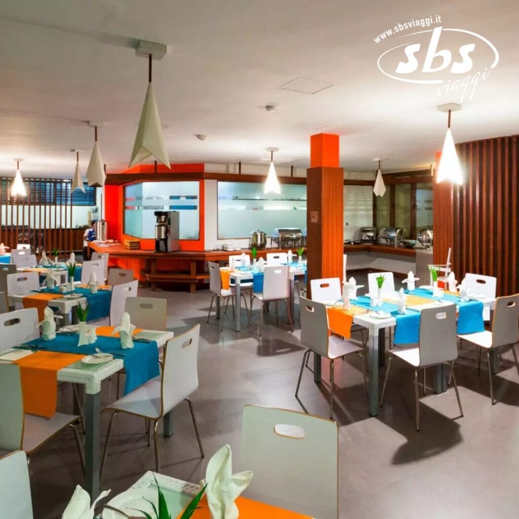Un moderno ristorante interno al Plumeria Hotel presenta coloratissime apparecchiature da tavola, con tovaglie arancioni e blu, sedie bianche e decorazioni di carta piegata. Il logo