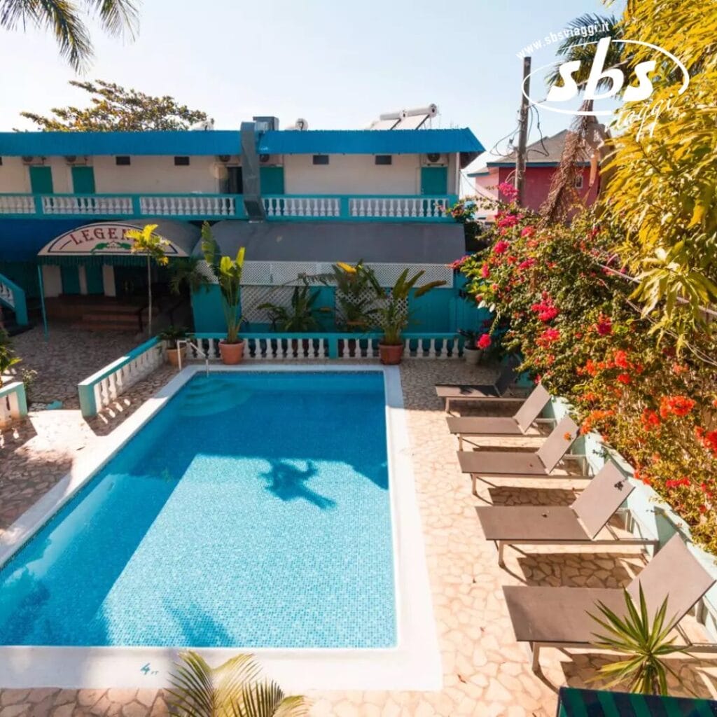 Una piscina scintilla di fronte a un edificio blu, impreziosita da sedie a sdraio su un patio in pietra e da piante tropicali che circondano l'area, creando un'invitante atmosfera di relax e tranquillità.