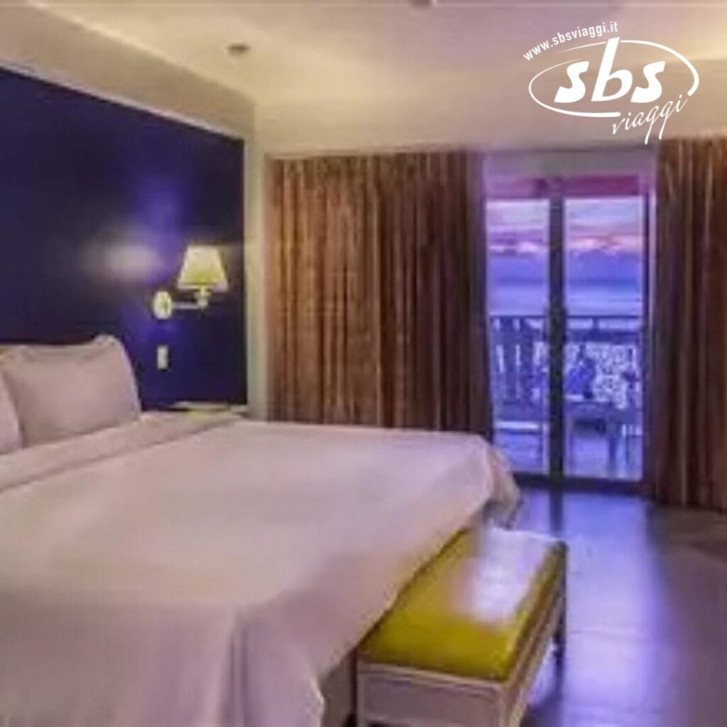 Camera d'albergo con un letto grande, lampada a parete e tende scure. Le porte del balcone rivelano una vista esterna sul Royal Decameron Montego Beach. Adatto a ospiti di età superiore ai 16 anni. Un logo SBS Viaggi è nell'angolo in alto a destra.