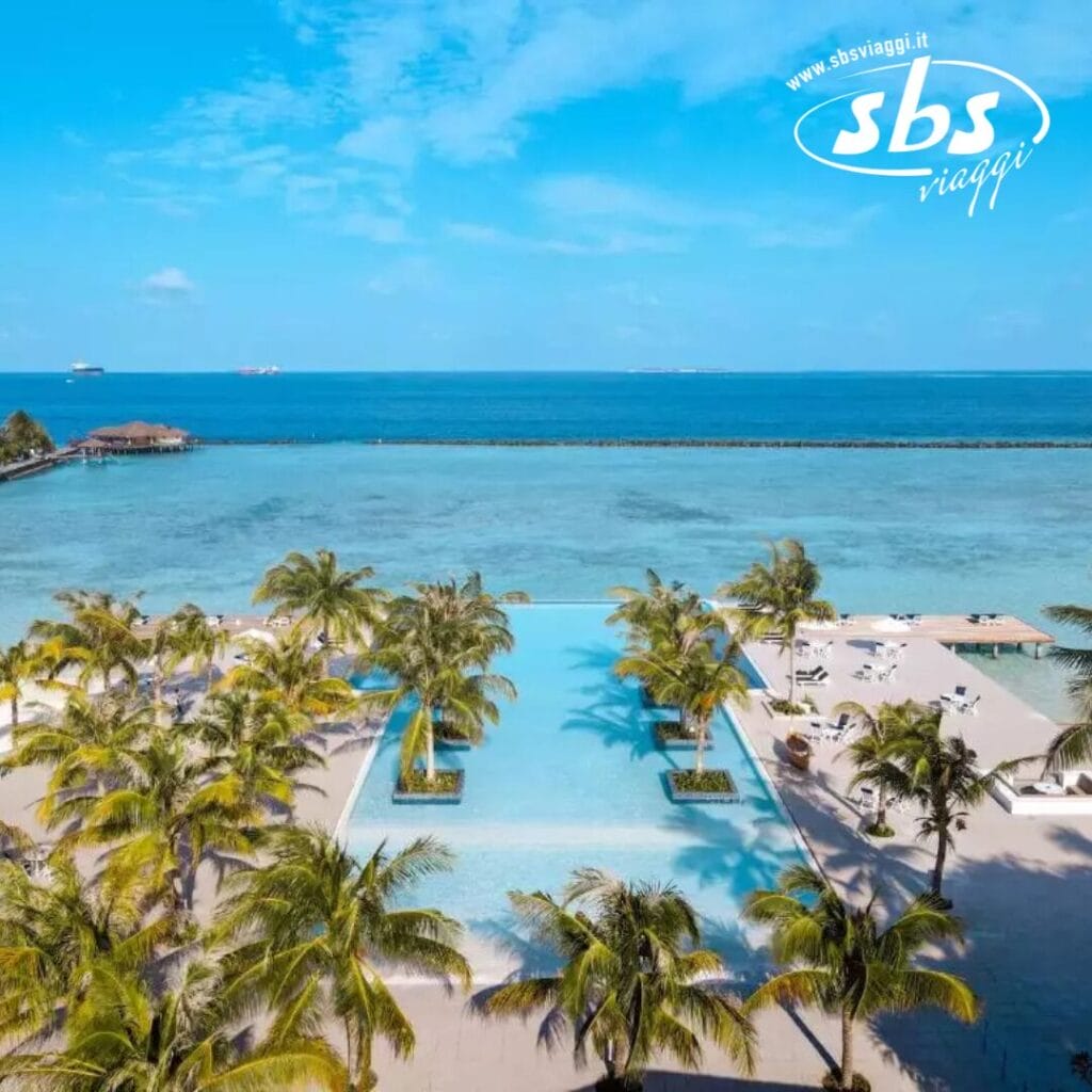 Una grande piscina a sfioro si affaccia su un limpido oceano azzurro, circondata da palme e lettini prendisole sotto un cielo luminoso, creando uno scenario che ricorda un'esplosione di paradiso.