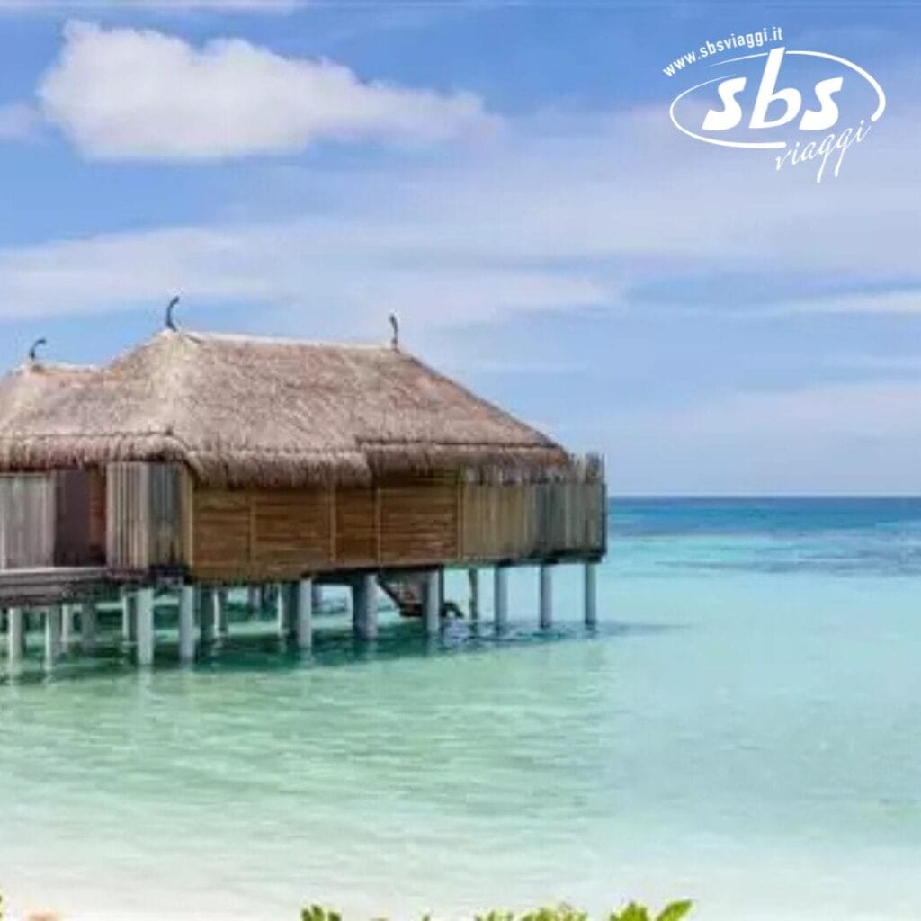 Una capanna con tetto di paglia su palafitte sopra le limpide acque turchesi del Constance Moofushi, Maldive, sotto un cielo parzialmente nuvoloso.