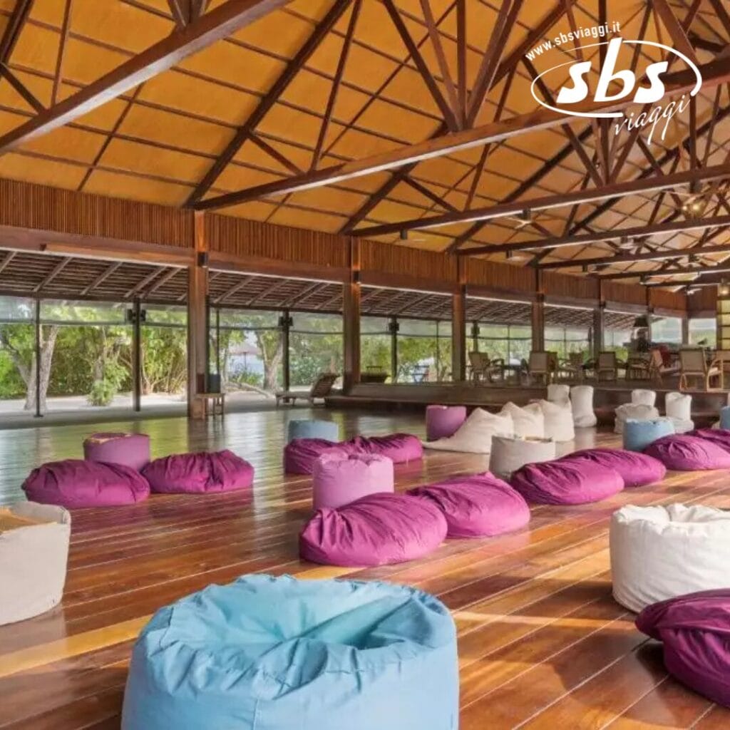 Una spaziosa stanza con pavimento in legno, ampie finestre e soffitto in legno, che ricorda il fascino naturale del Barefoot Eco Hotel. Sacchi di fagioli colorati sono sparsi in tutto lo spazio e il logo SBS Viaggi è visibile nell'angolo superiore.