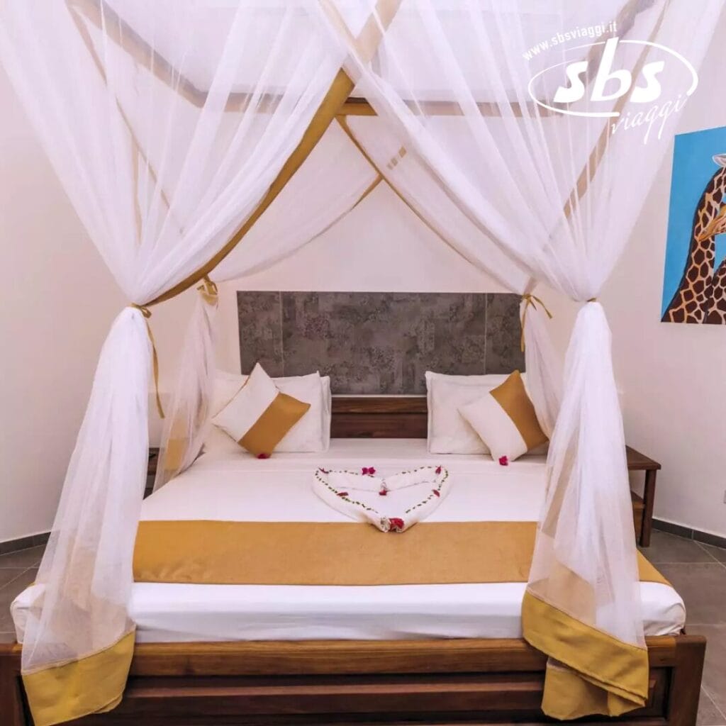 Un letto a baldacchino con lenzuola bianche e dorate, decorato con fiori disposti a forma di cuore, cattura l'essenza del lusso presso lo SBH Kilindini Resort. Un dipinto di giraffa è appeso elegantemente alla parete, aggiungendo un tocco di stravaganza a questo santuario in stile resort.