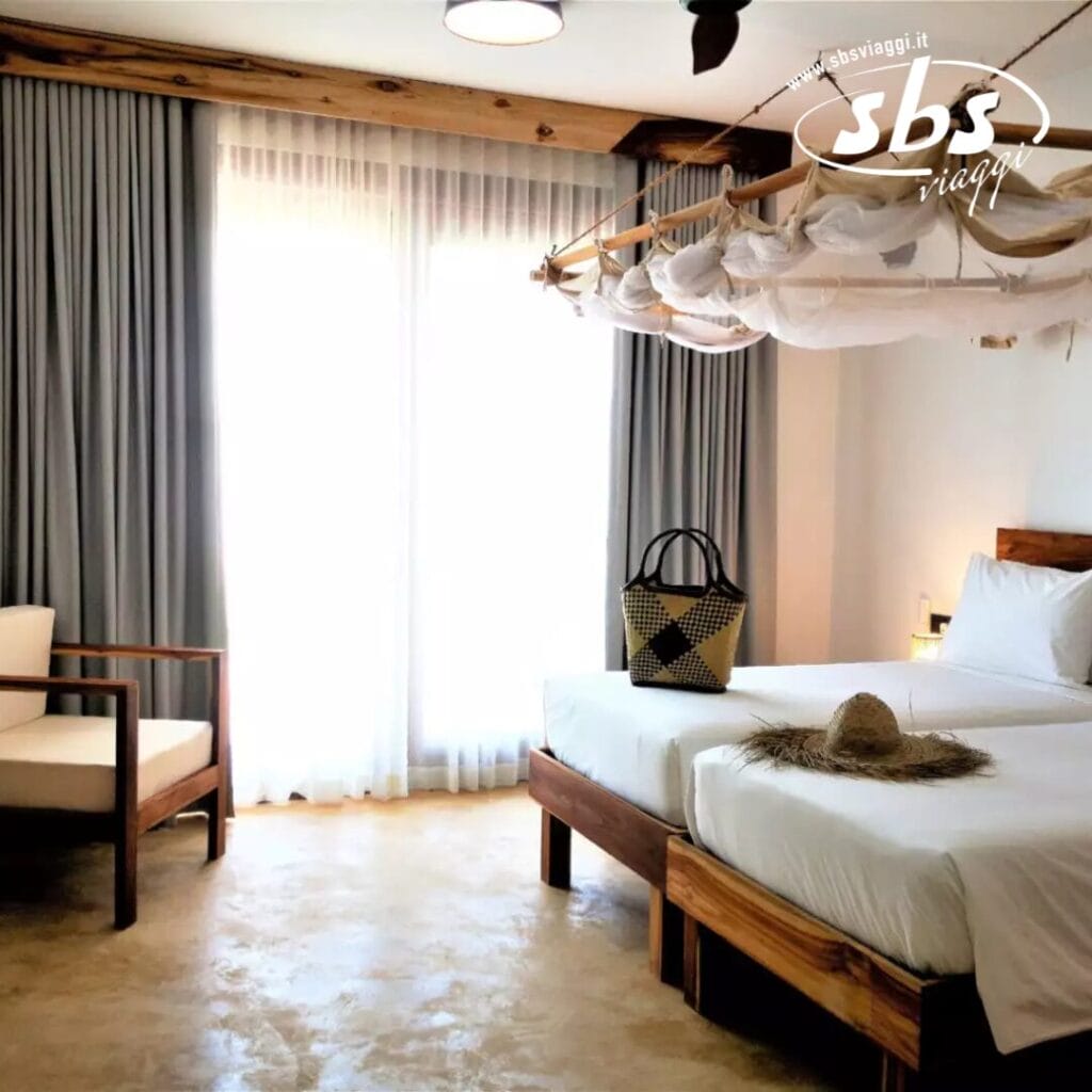 Una luminosa camera d'albergo presso lo SBH Monica Zanzibar Resort offre due letti, una sedia di legno e un cesto intrecciato. Tende trasparenti abbelliscono una grande finestra dove un cappello di paglia è appoggiato sul letto, sotto le travi a vista del soffitto.