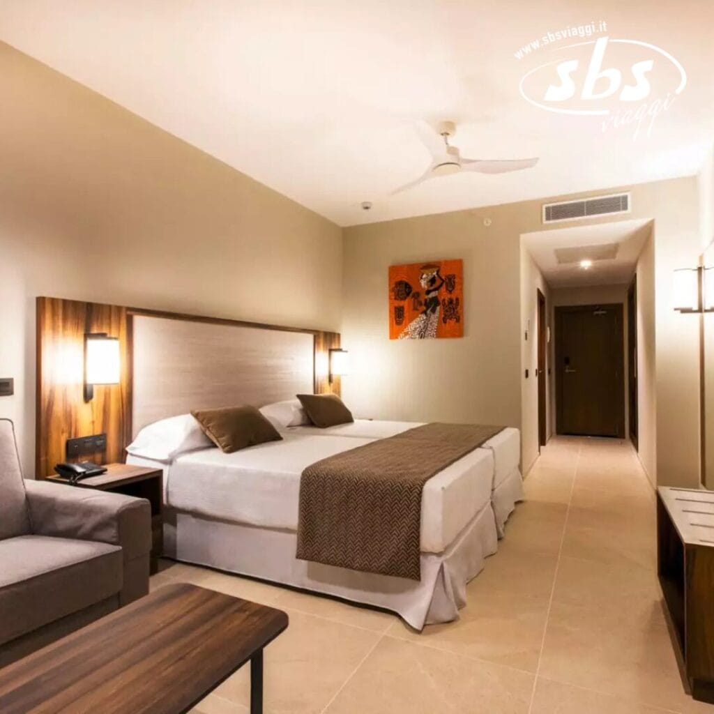 Al Riu Jambo Hotel, una camera moderna dispone di letti gemelli, un divano e un tavolo in legno. Luci calde e opere d'arte incorniciate adornano le pareti beige, mentre una porta conduce a un corridoio.