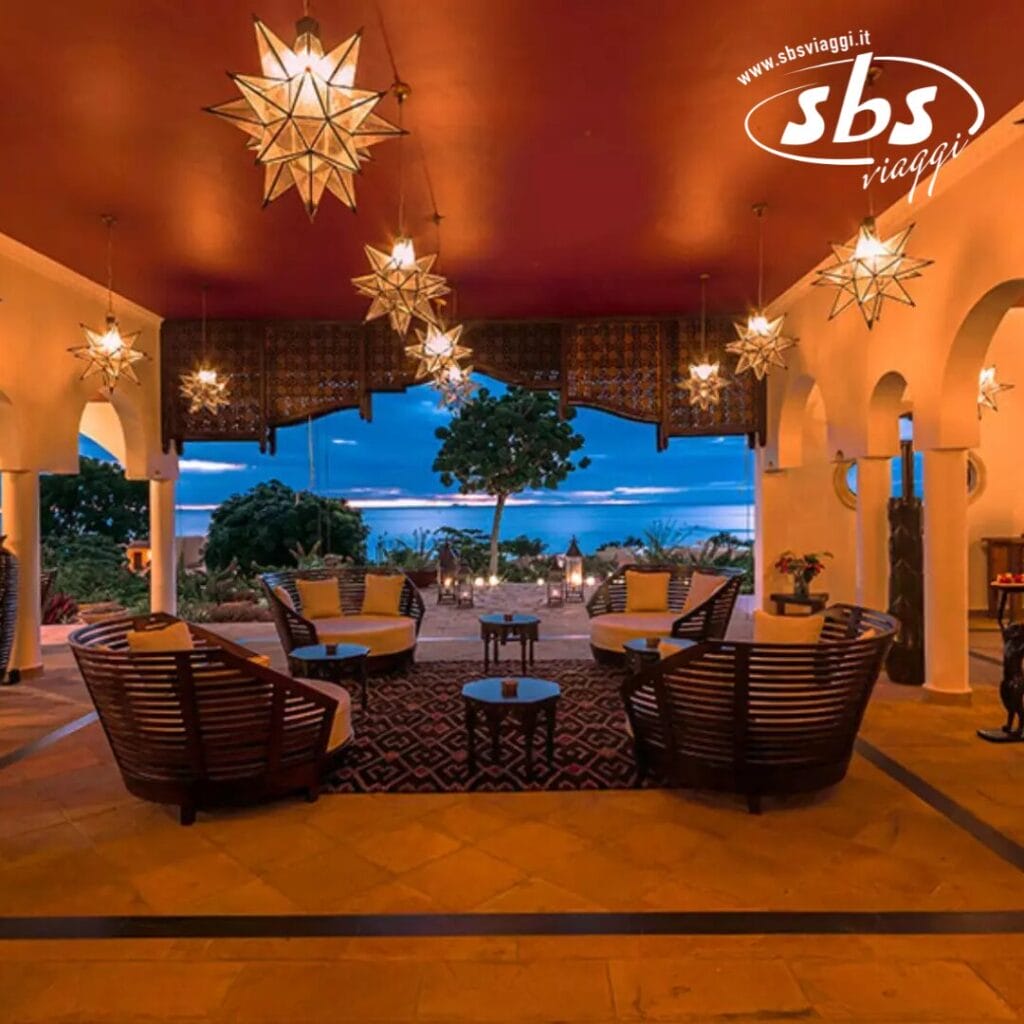 Una lussuosa area lounge esterna con posti a sedere in legno, lanterne decorative e una vista sull'oceano al tramonto cattura l'essenza di un resort di lusso. Il logo di SBS Viaggi adorna l'angolo in alto a destra, evocando l'eleganza del Riu Palace Zanzibar.
