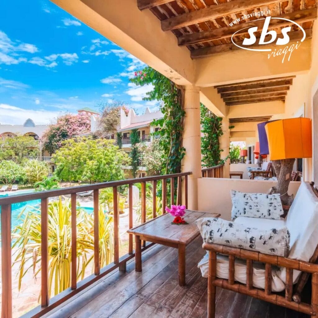 Un balcone in legno con posti a sedere al Lily Palm Beach Resort si affaccia su un giardino e una piscina sotto un cielo azzurro e limpido. Un logo con