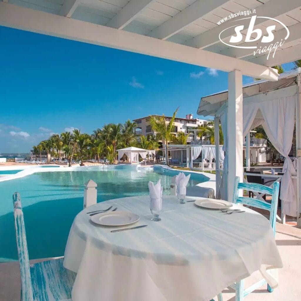 Un tavolo rotondo con una tovaglia bianca, apparecchiato per due, offre una vista incantevole sulla piscina del resort incorniciata da palme ed edifici che si stagliano contro un cielo azzurro e terso, creando lo sfondo perfetto per un'automatica atmosfera Bozza.