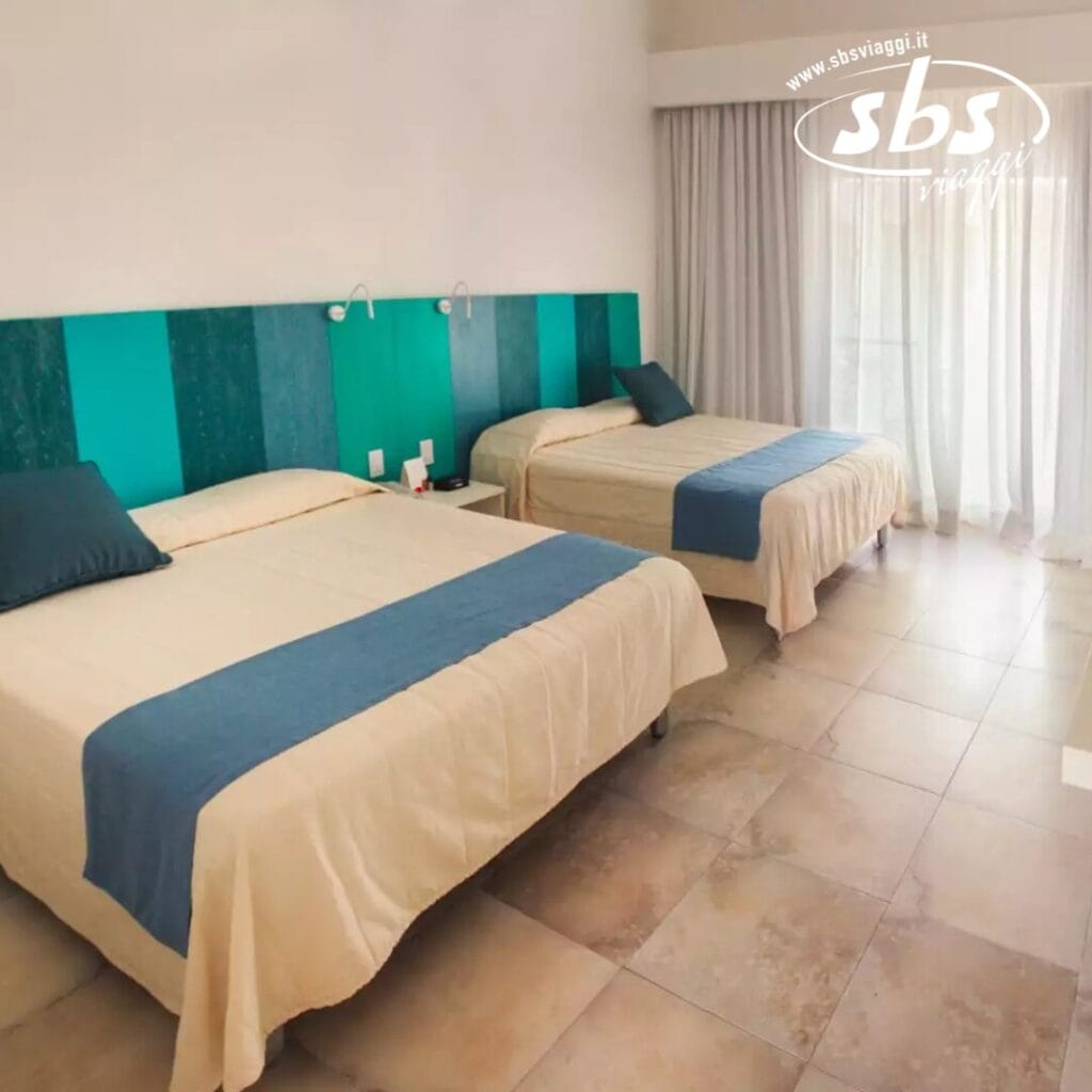 Due letti queen size con biancheria blu e beige decorano questa camera d'albergo con pavimento in piastrelle. Una porta a vetri sulla destra lascia entrare la luce naturale, evidenziando l'arredamento elegante. Nell'angolo in alto a destra, noterete il logo