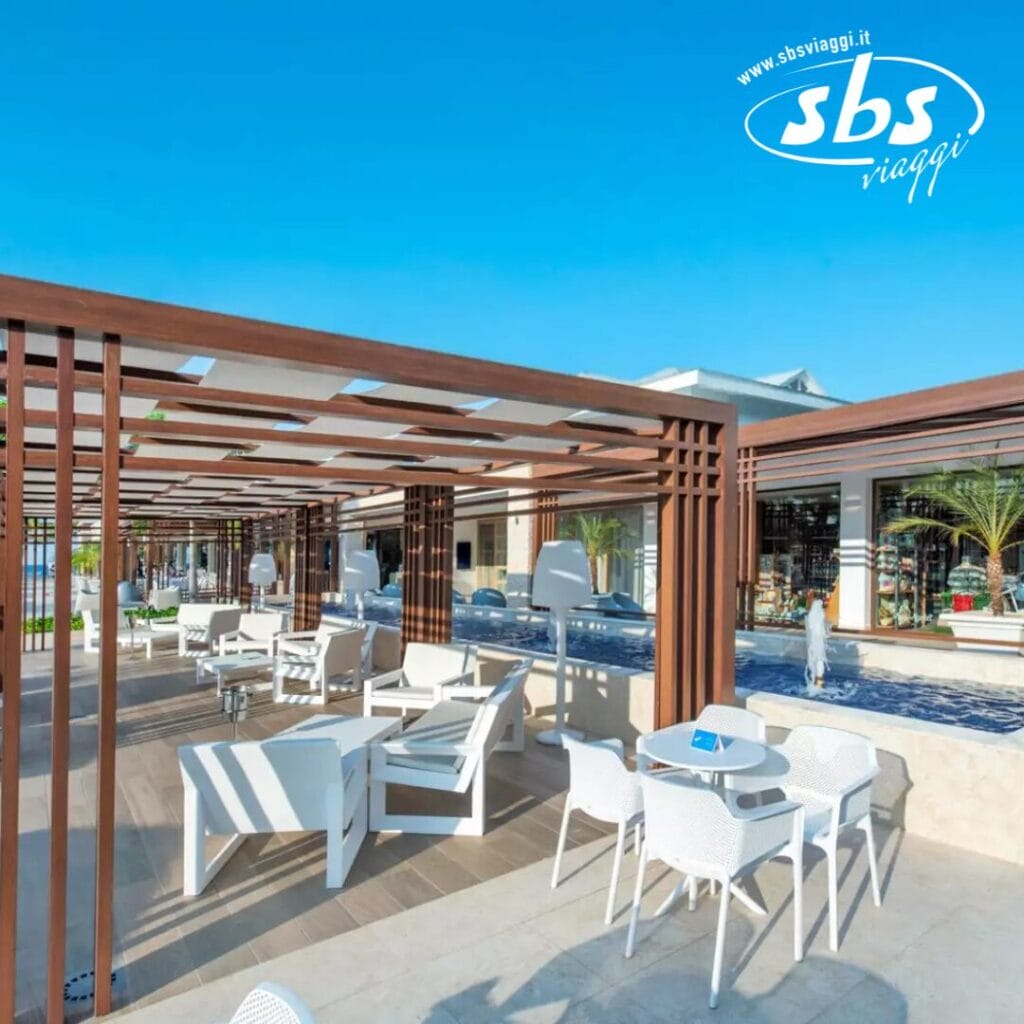 L'area salotto all'aperto è dotata di tavoli e sedie bianchi sotto pergolati in legno, con sfondo di un cielo azzurro e il logo