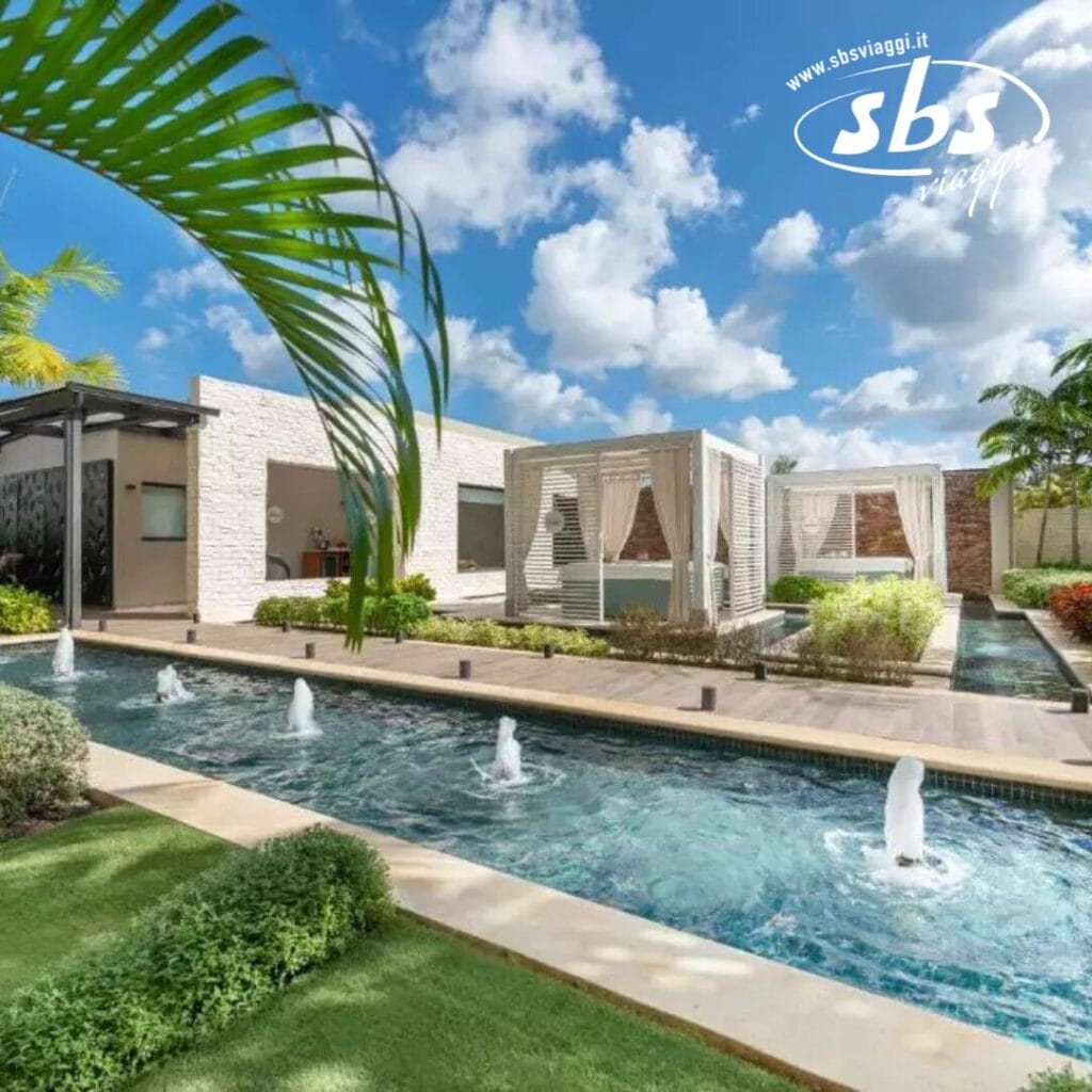 Lussuosa area esterna al Catalonia Royal La Romana, con piscina, fontane, cabanas e vegetazione lussureggiante sotto un cielo parzialmente nuvoloso. Progettata esclusivamente per gli over 18 che cercano un tranquillo relax.