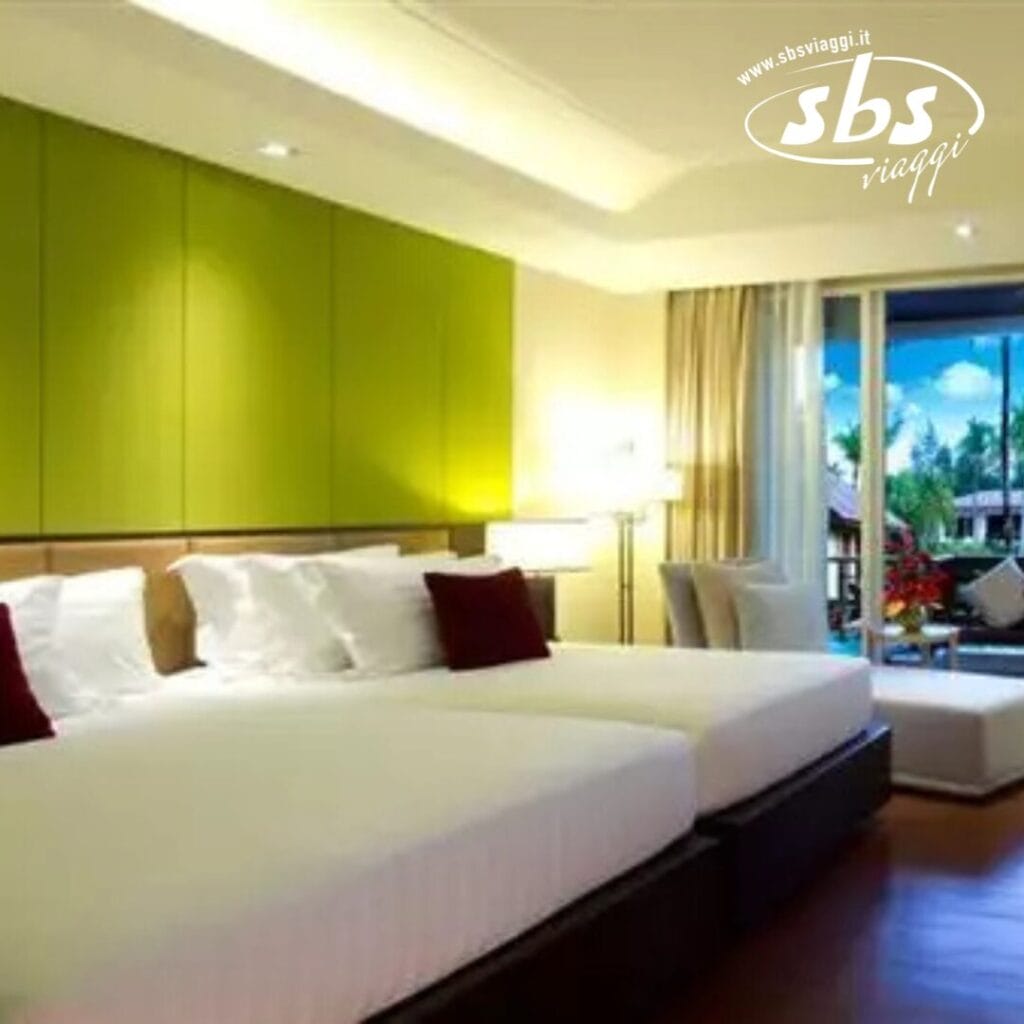 La camera dell'hotel resort è dotata di due letti, una parete verde e una poltrona. Le tende aperte rivelano una splendida vista sul giardino esterno, che richiama la tranquillità delle rinomate fughe del Kappa Club.