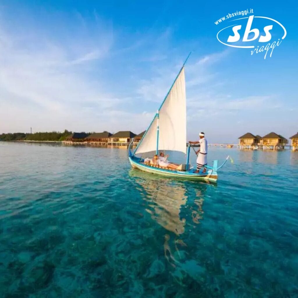 Una persona naviga su una piccola barca con una vela bianca su acque azzurre e limpide, godendosi il calore di un'isola estiva vicino ai bungalow sull'acqua sotto un cielo azzurro brillante.