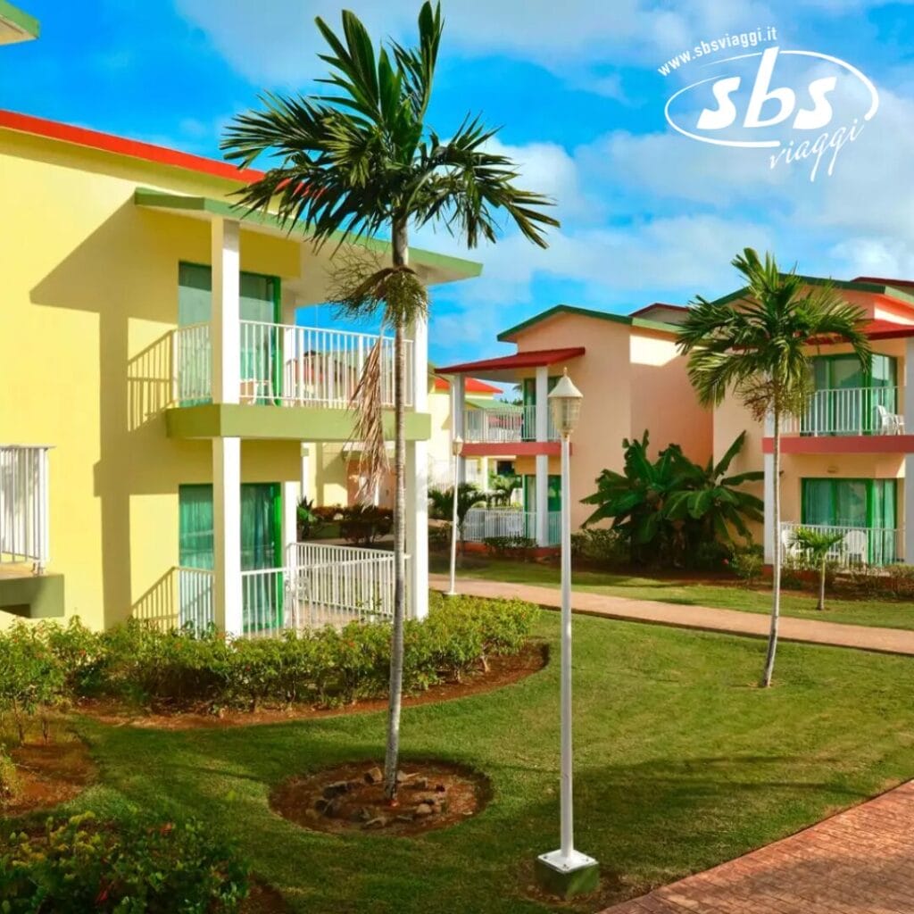 Gli edifici gialli a due piani con balconi verdi, incastonati tra palme e un prato ben curato sotto un cielo azzurro, presentano il caratteristico logo