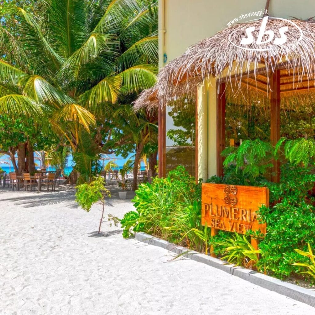 Un edificio fronte mare con tetto di paglia è circondato da palme e vegetazione lussureggiante. Un cartello recita con orgoglio