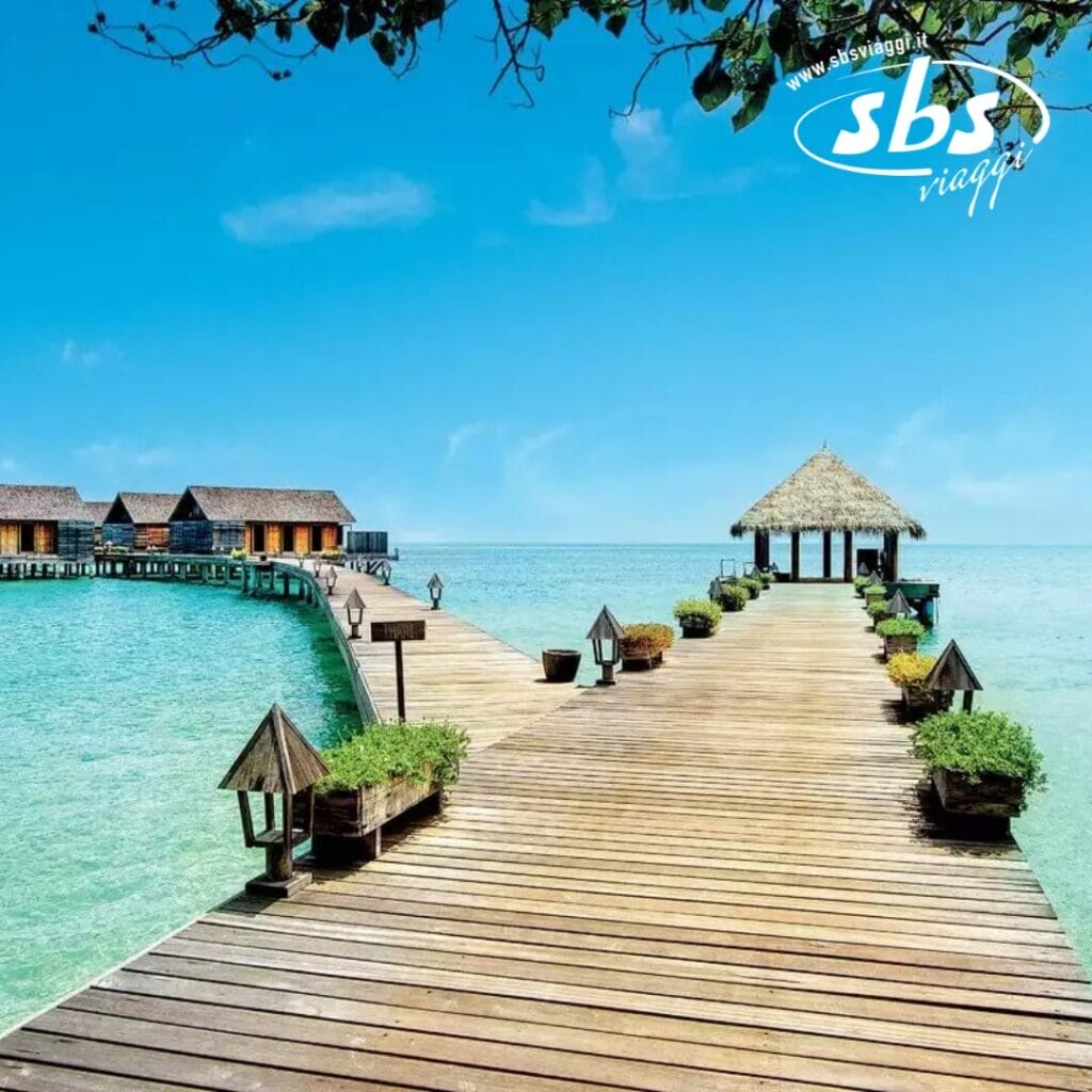 Una passerella di legno si estende su acque turchesi e trasparenti, conducendo alle ville con tetto di paglia del Gangehi Island Resort. Il cielo è terso e un gazebo sereno abbellisce il lato destro del sentiero, offrendo momenti di relax in spa.