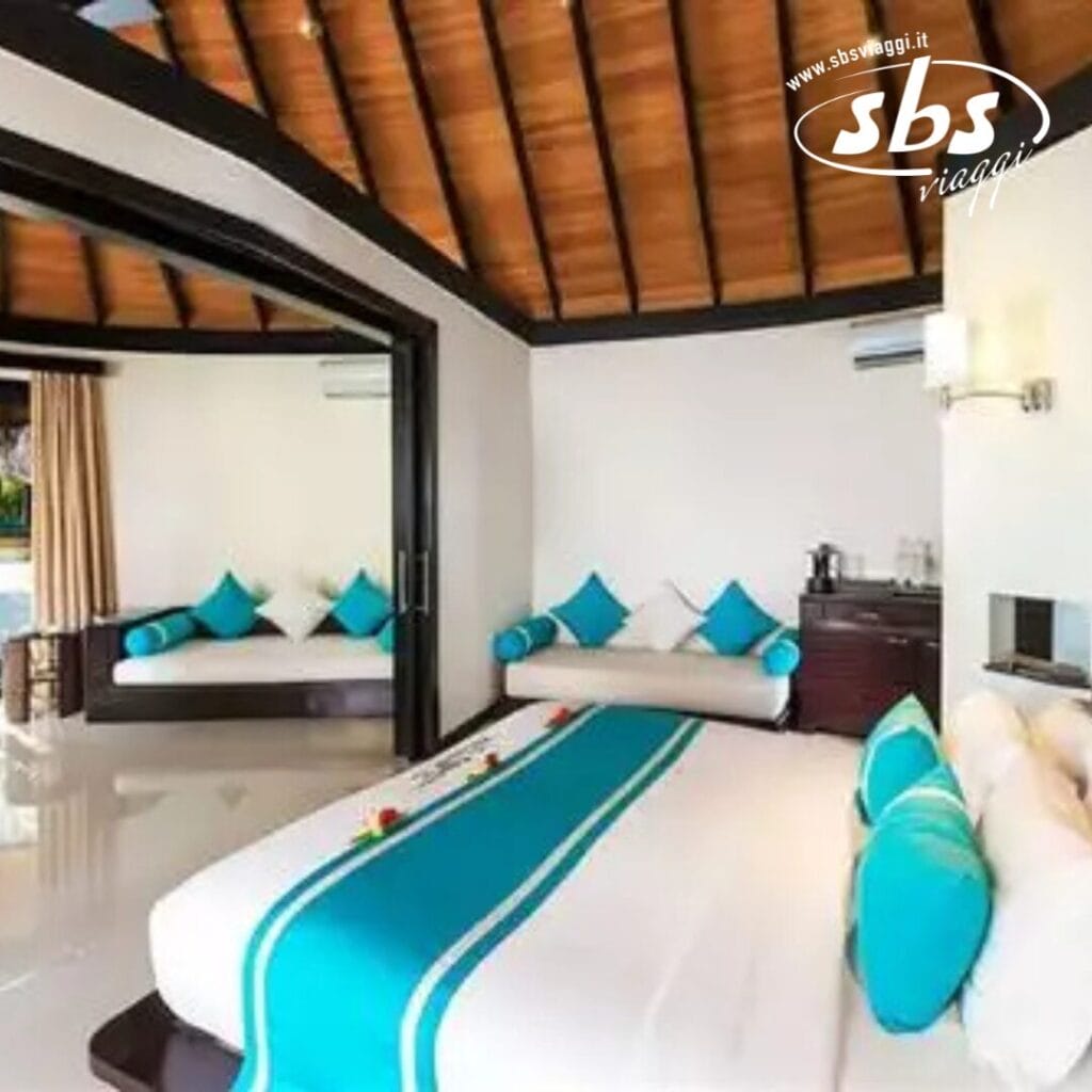 Una camera d'albergo al Sun Siyam Iru Fushi alle Maldive è dotata di un letto king-size decorato con lenzuola blu e bianche, un divano abbinato, uno specchio e un piccolo mobiletto. Il soffitto è impreziosito da eleganti travi in legno.