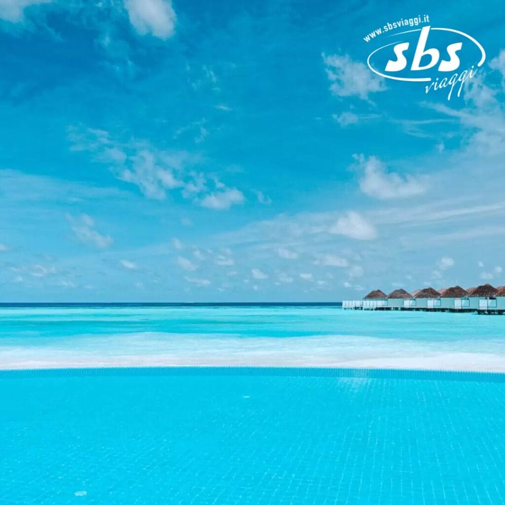 Una piscina a sfioro offre viste mozzafiato sull'oceano sotto un cielo azzurro con nuvole sparse, mentre i bungalow sull'acqua del Nova Maldives Resort punteggiano l'orizzonte, creando uno scenario pittoresco sull'isola di Vakarufalhi.