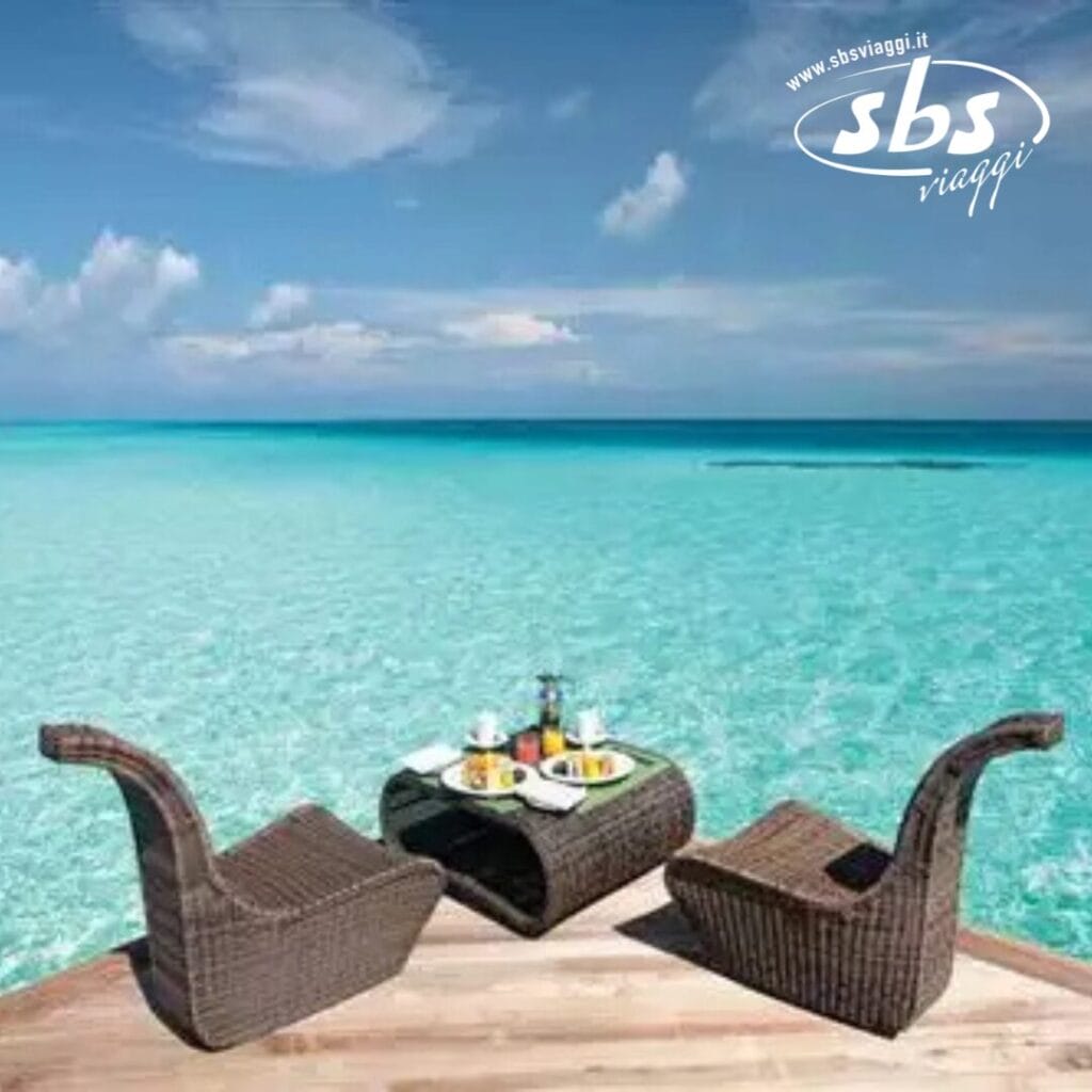 Due sedie in vimini e un tavolino con del cibo sono sistemati su una terrazza in legno, offrendo una vista serena sull'oceano turchese brillante del Constance Moofushi Maldives.