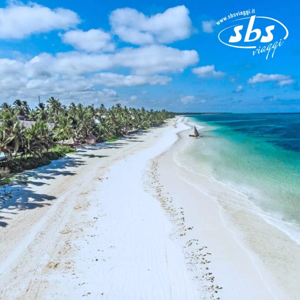Vista aerea di una spiaggia tropicale vicino al Sbh Kilindini Resort, con sabbia bianca, palme e acqua turchese. Una piccola barca a vela scivola vicino alla riva sotto un cielo parzialmente nuvoloso.