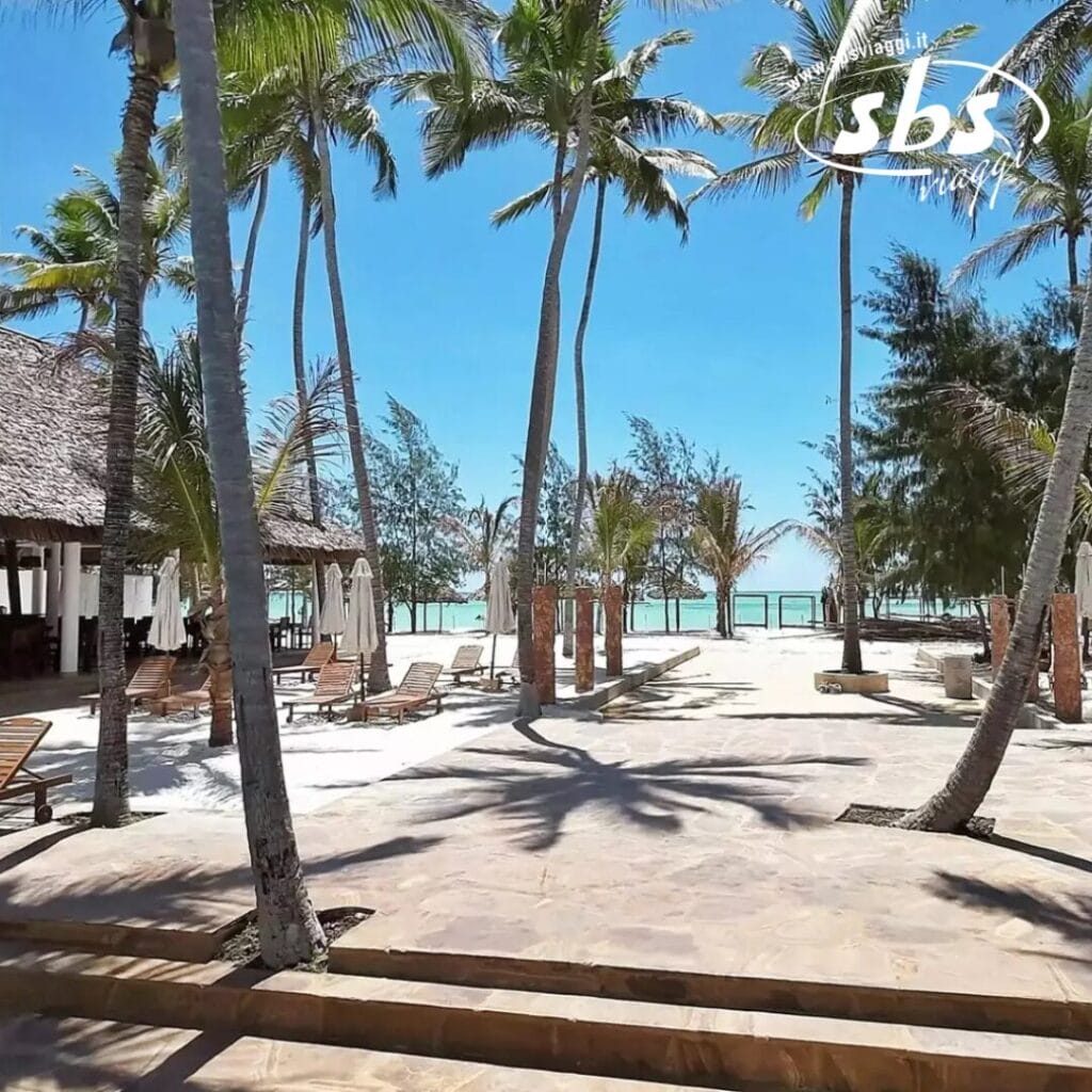 Una scena di spiaggia tropicale a Zanzibar presenta palme, lettini prendisole e un cielo azzurro e limpido. Una struttura con tetto di paglia è sulla sinistra e l'oceano luccica sullo sfondo, esaltando questo paradiso del resort.