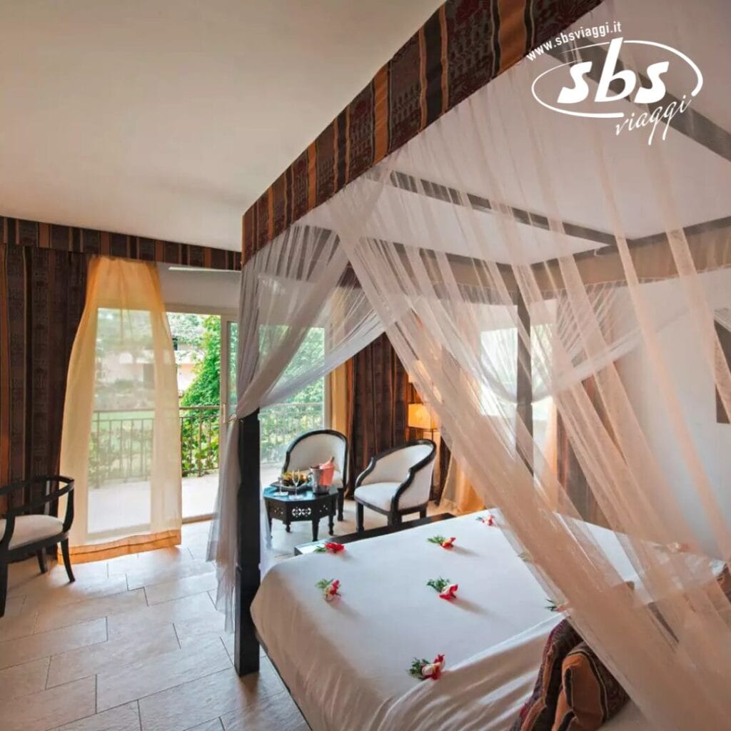 Una camera da letto da sogno con un letto a baldacchino decorato con fiori, due sedie vicino a una finestra e una vista sul verde esterno evoca una fuga serena che ricorda il Tui Blue Bahari.