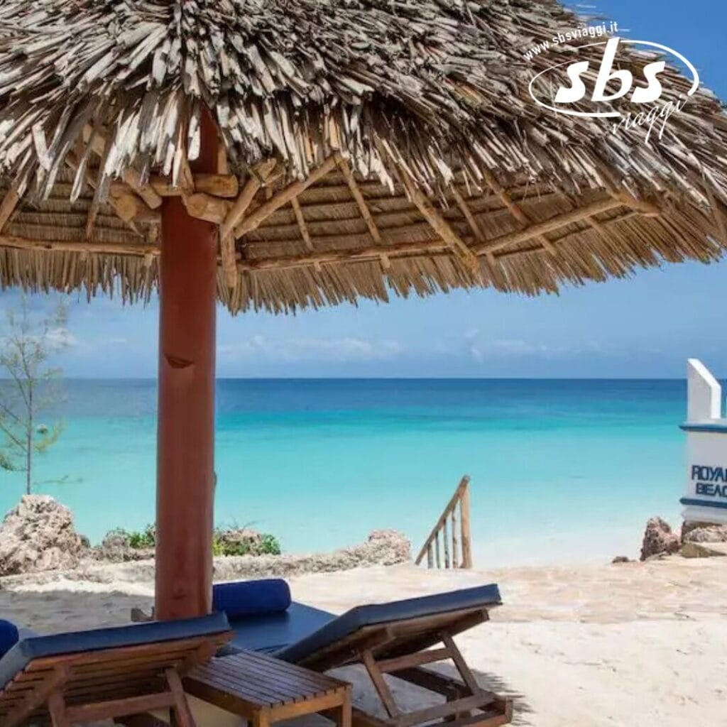 Scena di spiaggia al Royal Zanzibar con un ombrellone di paglia, sedie a sdraio e acqua cristallina sullo sfondo.