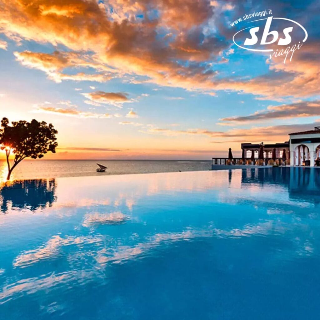 Piscina a sfioro al Riu Palace Zanzibar con vista sull'oceano, con cielo al tramonto, sagome di alberi e area lounge in lontananza. Logo SBS travel nell'angolo. Vivi l'atmosfera da hotel di lusso per eccellenza in questo incontaminato resort di Zanzibar.