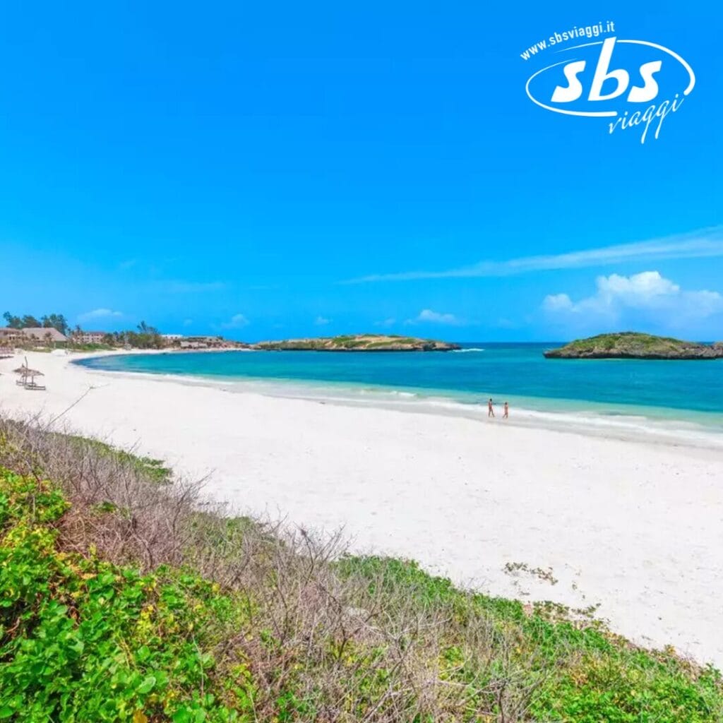 Spiaggia sabbiosa con acqua azzurra e limpida sotto un cielo luminoso. Due persone passeggiano lungo la riva, incorniciate da una vegetazione lussureggiante in primo piano. Scopri la tranquillità al Barracuda Inn, dove il paradiso incontra il comfort. Logo SBS Viaggi nell'angolo in alto a destra.