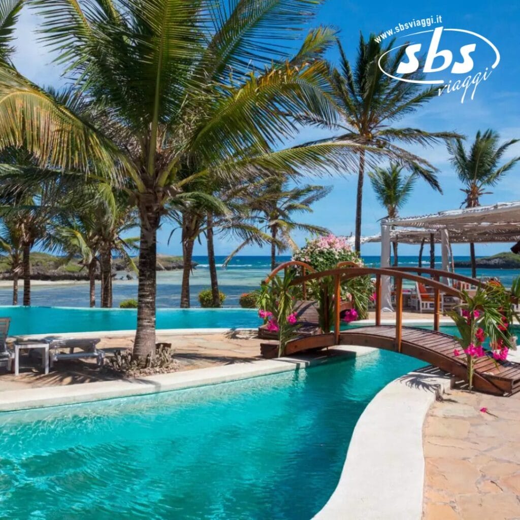 La serena scena tropicale del Lily Palm Beach Resort presenta una piscina tranquilla con palme che ondeggiano dolcemente. Un affascinante ponte di legno si inarca con grazia sull'acqua e comode sedie a sdraio invitano sul ponte di pietra, il tutto sullo sfondo dell'oceano scintillante e di un cielo azzurro limpido.