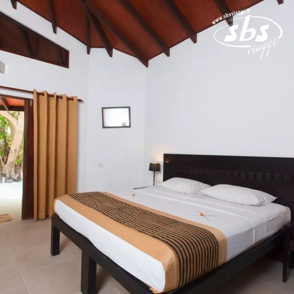 Una serena camera da letto d'albergo all'Embudu Village, dotata di un letto matrimoniale con lenzuola marroni e bianche, lampade da comodino e travi in legno rustico sul soffitto. Una tenda vela parzialmente la porta, aggiungendo privacy a questo ambiente da resort sull'isola, mentre un piccolo specchio a parete completa il fascino.