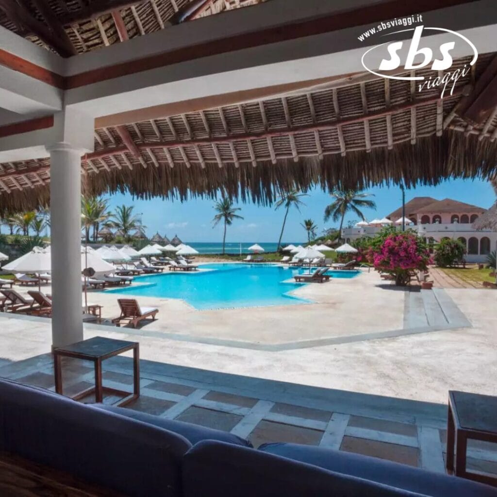 Vista della piscina di un resort con sedie a sdraio, ombrelloni e un tetto di paglia che si affaccia sull'oceano e sulle palme sotto un cielo azzurro e limpido.