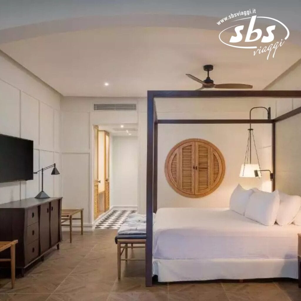 La camera d'albergo è dotata di un grazioso letto a baldacchino, una TV a parete, una cassettiera in legno e un ventilatore a soffitto. Una decorazione circolare in legno rustico aggiunge carattere, mentre una porta conduce a un'altra stanza. Bozza automatica suggerisce opzioni aggiuntive per ottimizzare la tua esperienza di soggiorno con tocchi personalizzati.