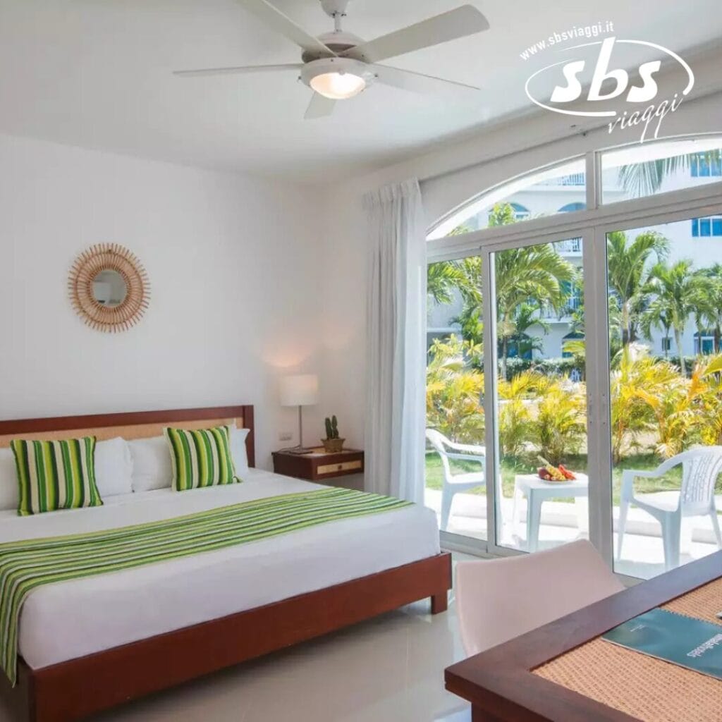 Una luminosa camera d'albergo con un letto king-size, cuscini a strisce verdi e una vista sul giardino attraverso grandi porte a vetri. Le pareti bianche, il ventilatore a soffitto e i semplici mobili in legno creano un'atmosfera invitante. Per approfondimenti di settore o specifiche del pubblico di destinazione, potrebbero essere necessari maggiori dettagli per un'integrazione SEO ottimale.