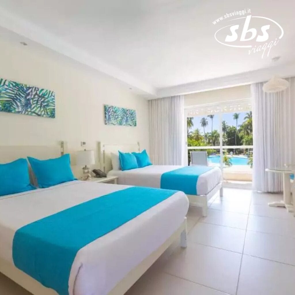 Camera d'albergo con due letti con accenti turchesi, un tavolino e una vista sulla piscina e sul giardino tropicale attraverso grandi finestre nel nostro resort sulla spiaggia di Punta Cana. Godetevi l'accesso alla nostra lussuosa spa per un soggiorno davvero rilassante.