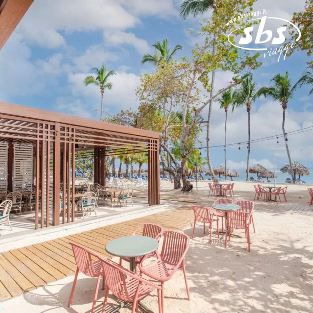 Ristorante all'aperto sulla spiaggia al Grand Bahia Principe con sedie e tavoli rosa sulla sabbia, sotto un pergolato di legno. Nelle vicinanze, palme e ombrelloni di paglia costeggiano una spiaggia sabbiosa di El Portillo. Il cielo è soleggiato con nuvole sparse.