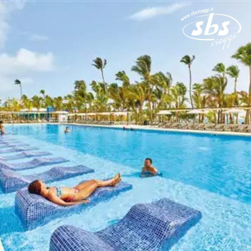 Persone che si rilassano sui lettini della grande piscina all'aperto del Riu Palace Punta Cana, con le palme che ondeggiano sullo sfondo in una giornata di sole.