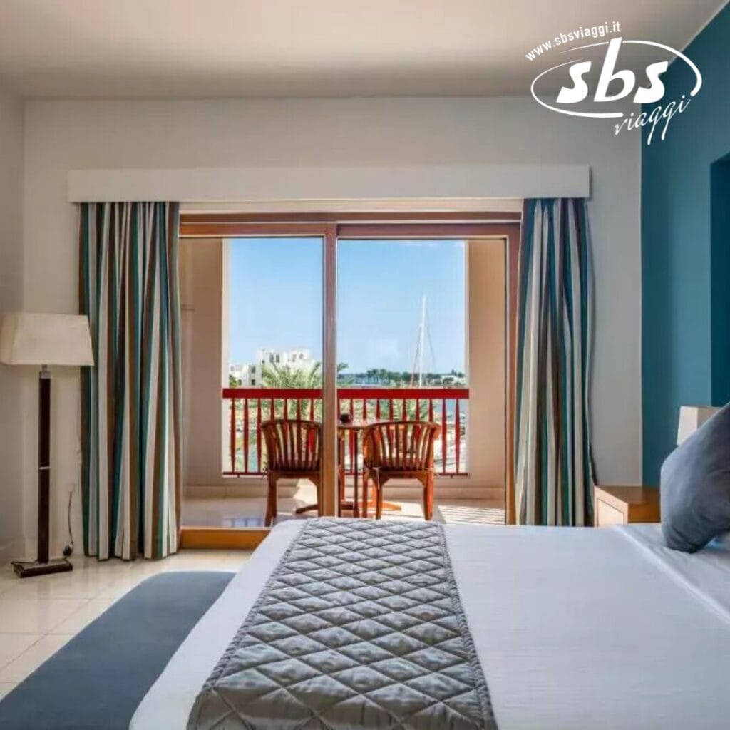 Interno della camera d'albergo con un letto ben fatto al Sifawy Boutique Hotel, accanto a una grande finestra. Fuori, c'è un balcone con due sedie e una vista di barche a vela contro un cielo limpido. Logo del Kappa Club nell'angolo.