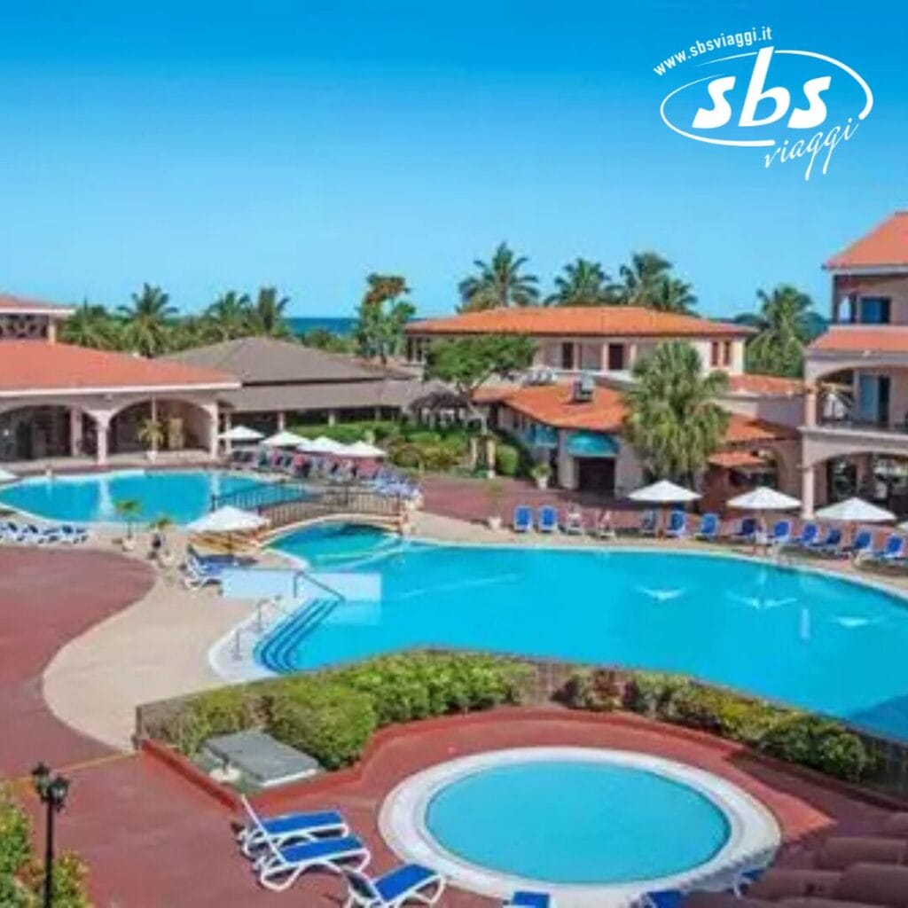 Un resort con numerose piscine all'aperto, circondato da sedie a sdraio, palme verdi e edifici con tetti in terracotta sotto un cielo azzurro e terso, offre una fuga idilliaca. Provate il massimo relax in questo rifugio Bozza automatica.