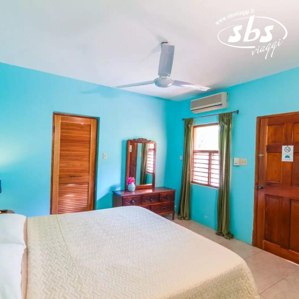 Una camera da letto con pareti blu, un ventilatore a soffitto, un letto e una cassettiera con uno specchio vi danno il benvenuto al Sunrise Club. La porta di legno si apre su una finestra con tende verdi che svolazzano nella brezza dell'unità di aria condizionata.