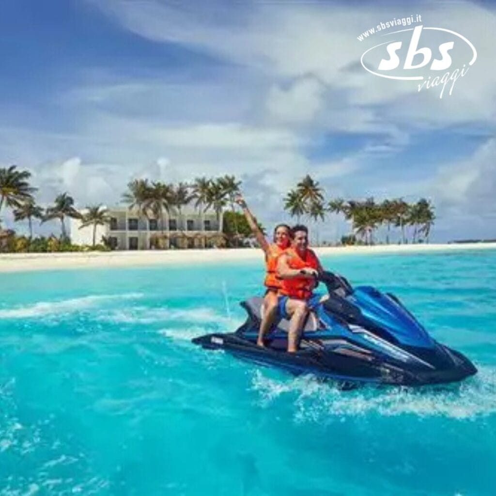 Due persone stanno guidando una moto d'acqua sulle acque cristalline vicino a un'isola tropicale delle Maldive, con l'iconico edificio bianco e le palme dell'atollo Riu che impreziosiscono lo sfondo.