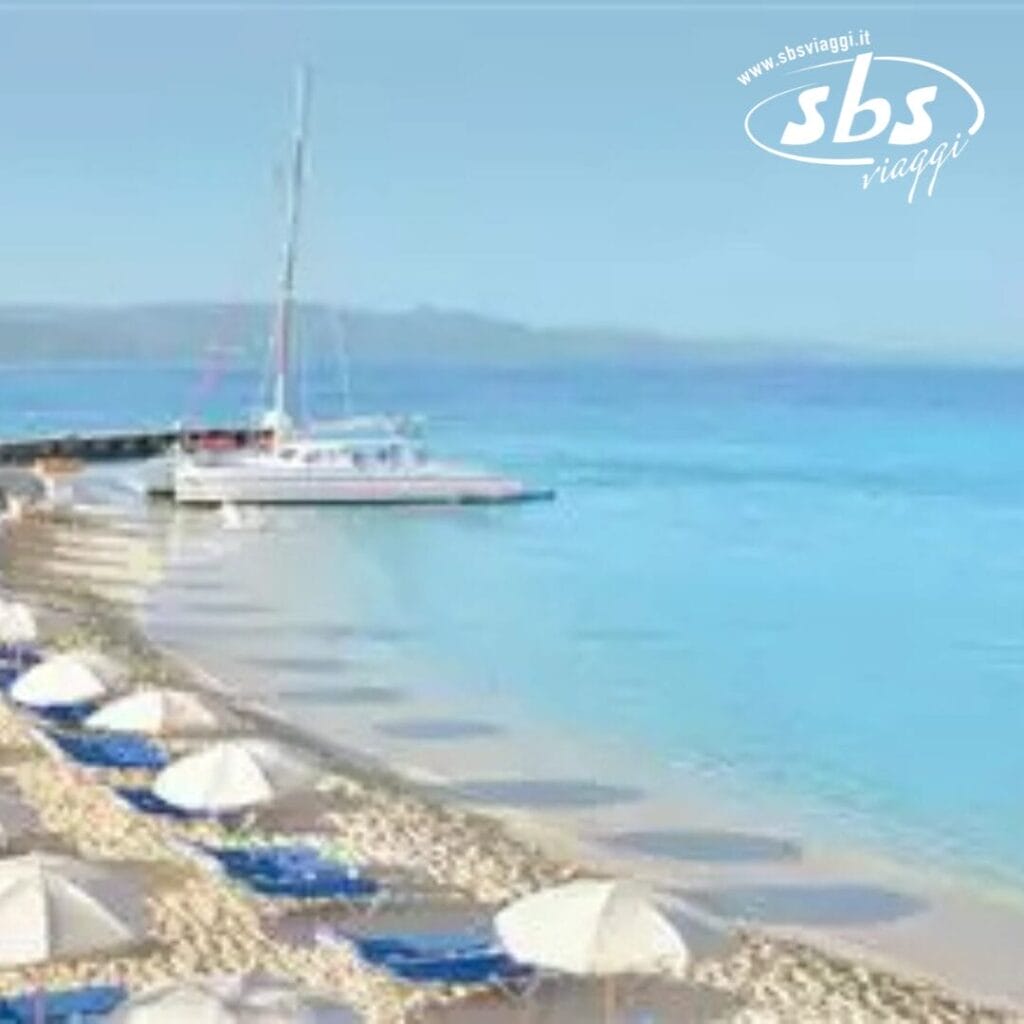 Un catamarano riposa sulle rive sabbiose del Royal Decameron Montego Beach, circondato da ombrelloni e sedie a sdraio blu. L'acqua cristallina luccica sotto un cielo azzurro. Perfetto per gli ospiti di età superiore ai 16 anni in cerca di tranquillità. Logo nell'angolo in alto a destra.